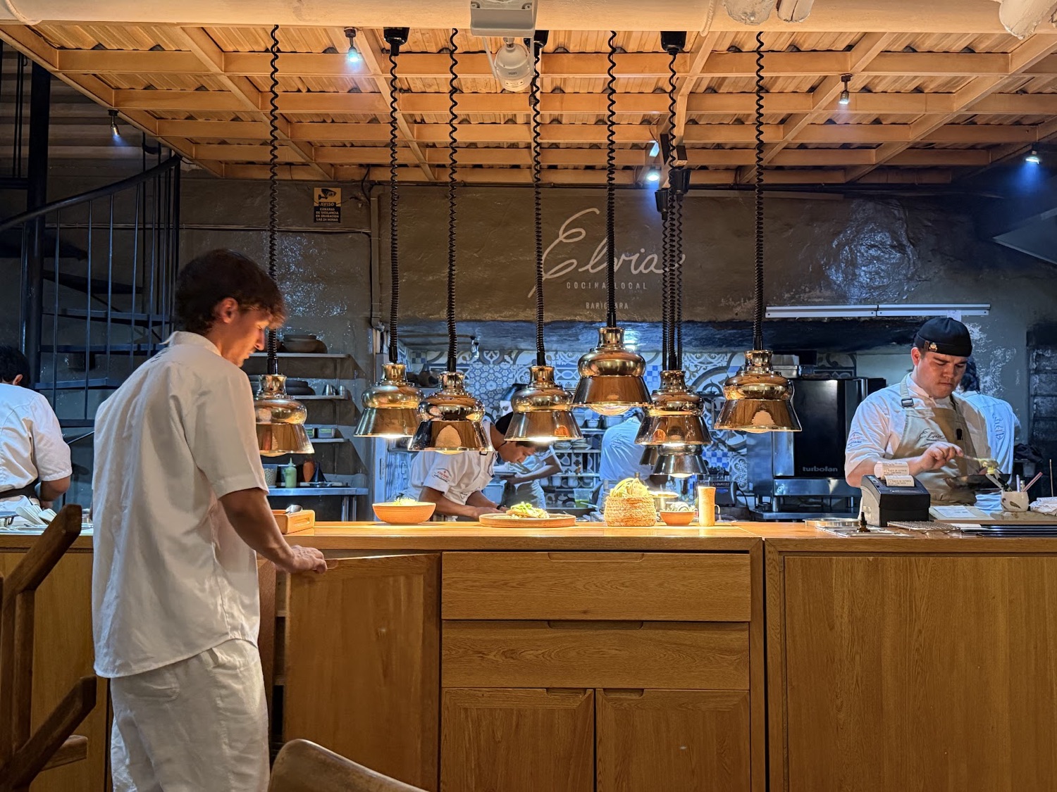 Interior de Elvia: barra de madera, lámparas de cobre y cocineros trabajando