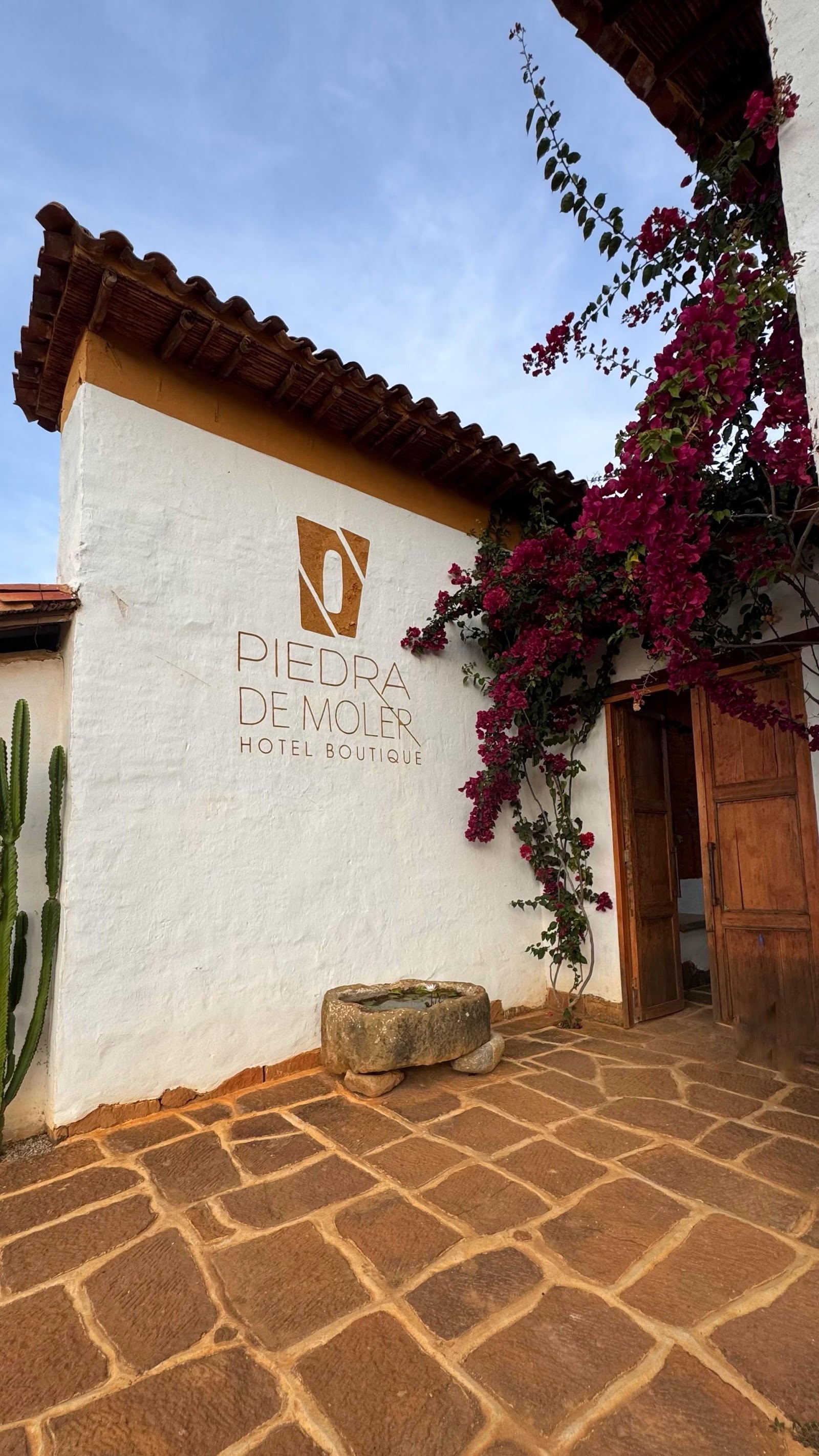 La entrada del hotel con su logo pintado en la pared y buganvilias